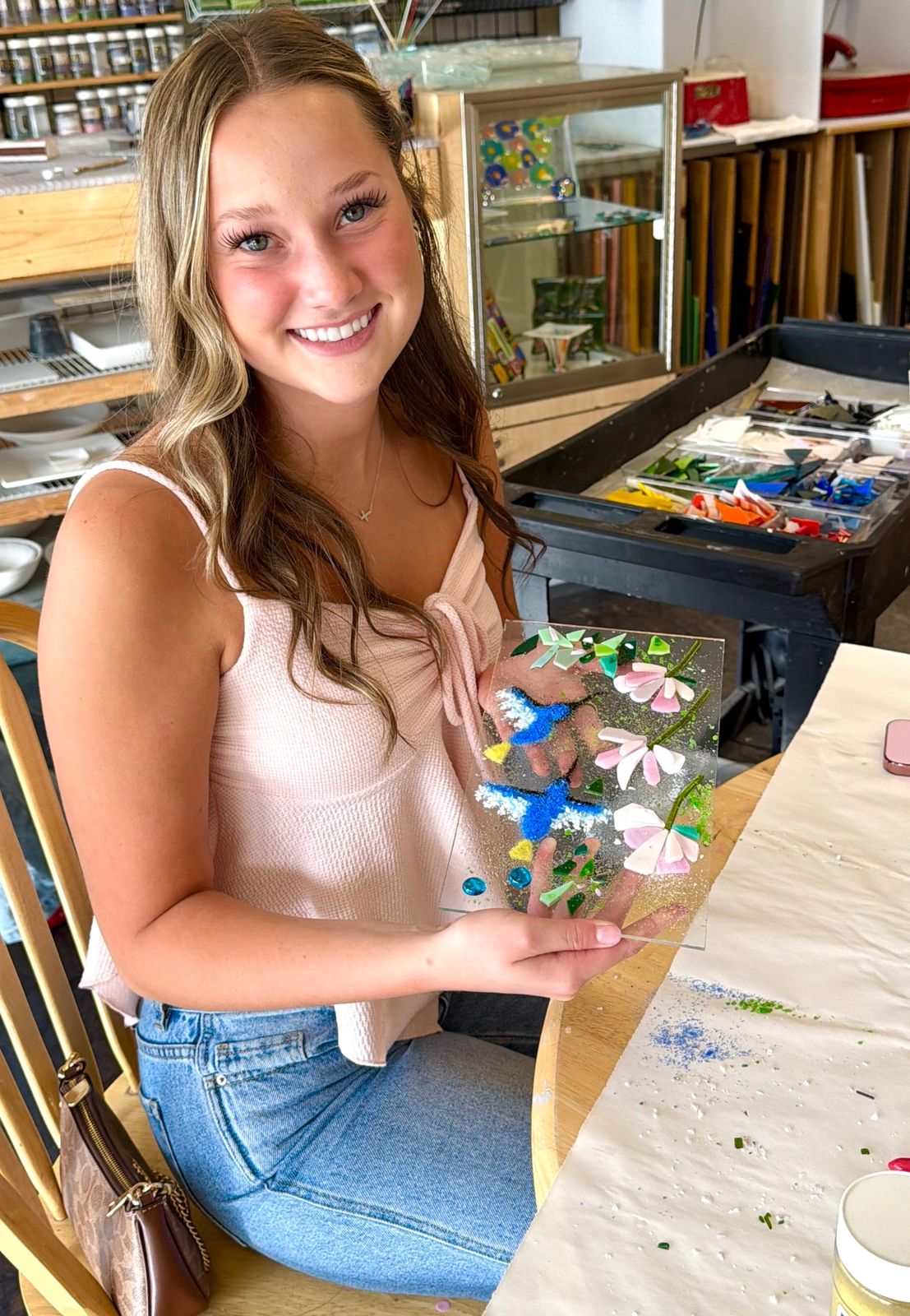 A smiling first-time glass fuser holding her finished hummingbird and flowers glass panel at Paint Fusion Park City