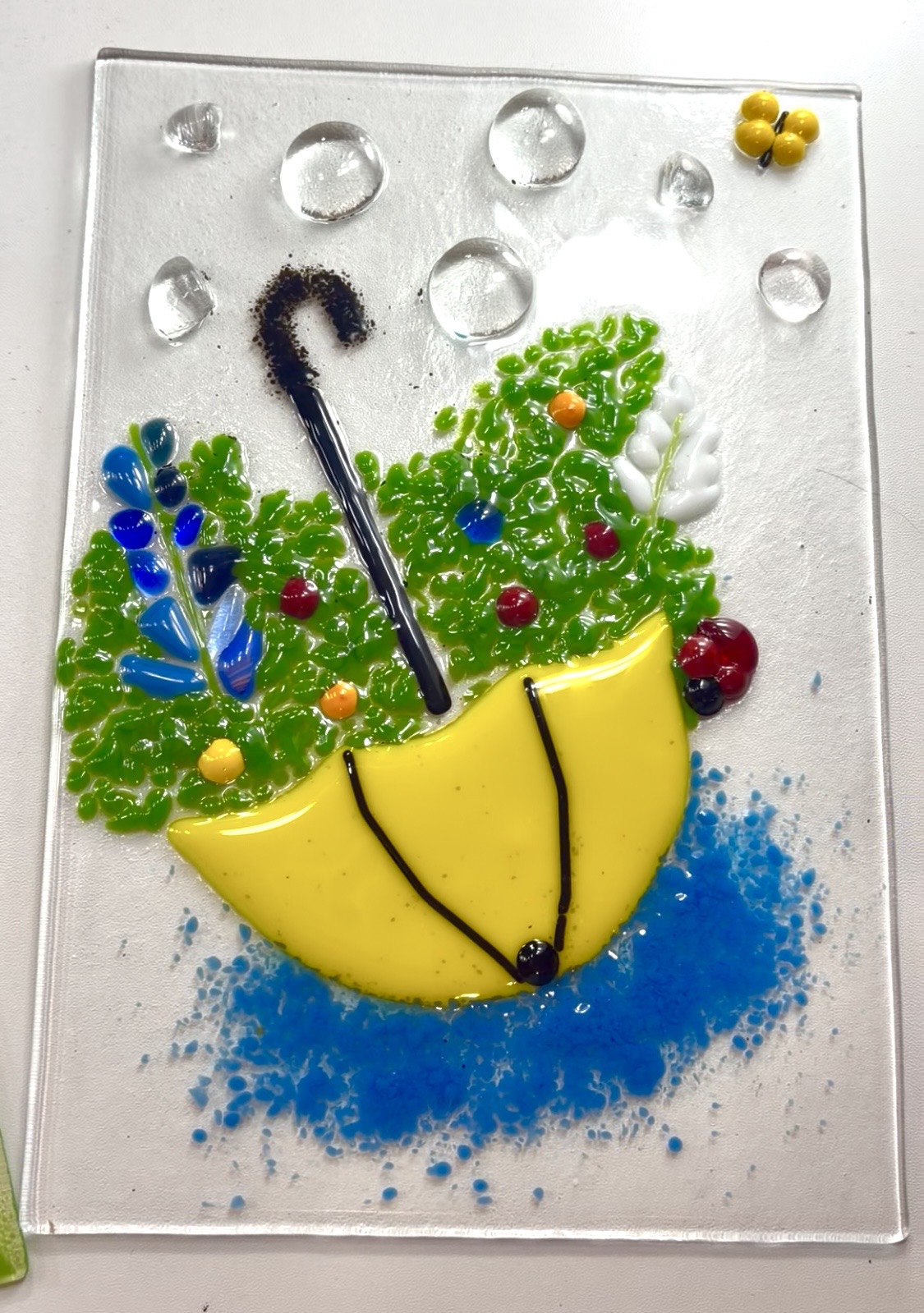 A fused glass panel of a yellow umbrella and flowers made at Paint Fusion Park City